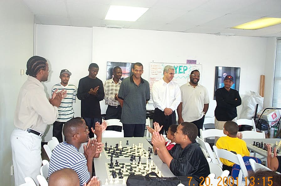  GM Maurice Ashley (standing left) introduces Wilbert Paige participants to Harlem Chess Center students. The players are: (L-R) FM Kenny Solomon, IM Amon Simutowe, NM Grace Nsubuga, FM Stephen Muhammad, NM Ernest Colding, NM Norman Rogers, and IM Watu Kobese. Copyright  2001, Daaim Shabazz.