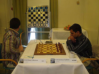 Hikaru Nakamura appears to glance at 2.Qh5!? on the demo board as Krishnan Sasikiran ponders. Nakamura got the idea to play the move from Jason Doss druing a chat on the ICC. Doss is one of Parham's former students, but his urging was initially a light-hearted joke. What was initially a joke turned into reality when Nakamura played 2.Qh5.  Photo by Sigeman & Co., 2005.