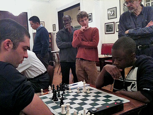 Joshua Colas (right) in action versus NM Leonardo Martinez at the Marshall Chess Club. Guy Colas in background watching. Photo by James Black, Sr.