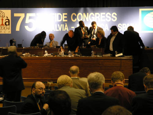 FIDE General Assembly during an intermission. On the stage at the far left is Sri Lankas Derrick Perera (FIDEs General Secretary of Asia) conferring with former FIDE President Florencio Campomanes. Standing in the middle is Jamaica delegate Ian Wilkinson and Barbados delegate Allan Herbert making a point about FIDE federation dues to FIDE official. FIDEs continental President Nizar ElHaj of Libya is to the right of that group. ElHaj had complained earlier because the organizing committee did not have placards for the African federations who ended up seated in the rear.