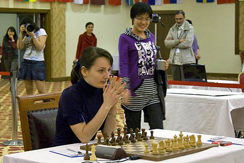 GM Kateryna Lahno waits patiently as a very cheerful Hou Yifan arrives at the board. Very interested spectators in background including Koneru Ashok, father of Koneru Humpy and Wang Qian, mother of Hou Yifan. Photo by wwcxc2010.tsf.org.tr.